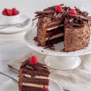 A chocolate layer cake on a white cake stand with one slice cut and plated